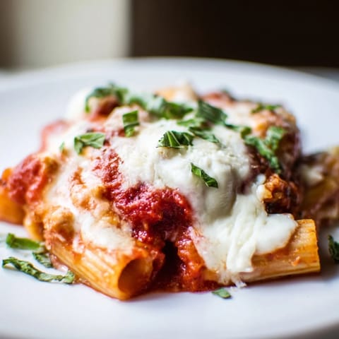 Melting mozzarella covers the Cheesy Baked Ziti, a comforting casserole with basil garnish, prepared fresh.