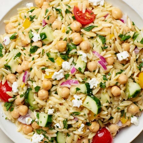A close-up of the lemony chickpea orzo salad, featuring fresh parsley and mint leaves, juicy tomato halves, and crisp diced cucumbers for a refreshing Mediterranean side.